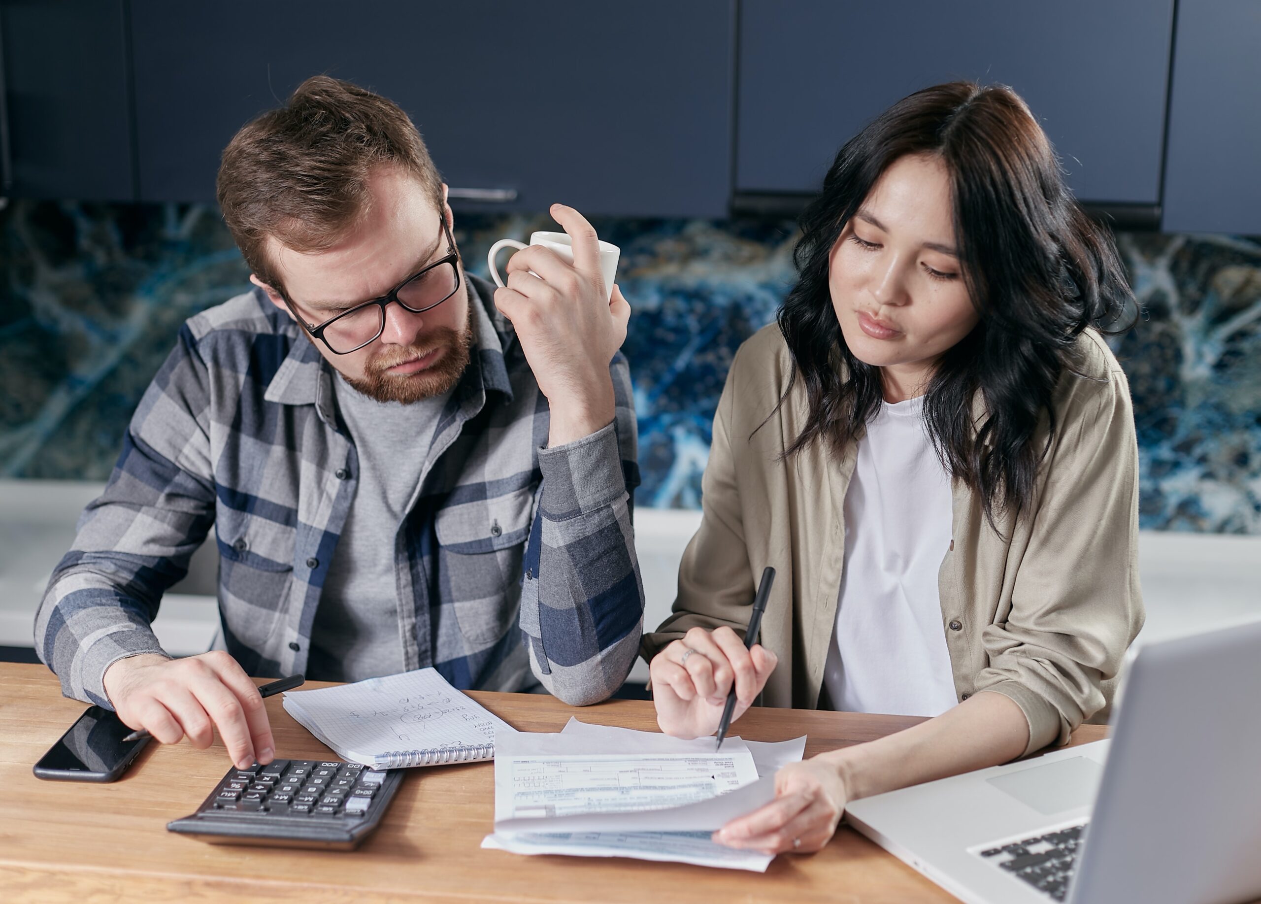 Casal sentado à mesa da cozinha revisando contas com calculadora, papéis e notebook